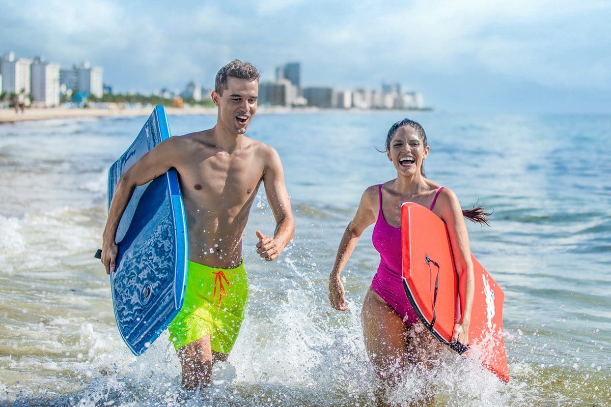 Couple running on the beach with bodyboards in Miami - Visit Florida
